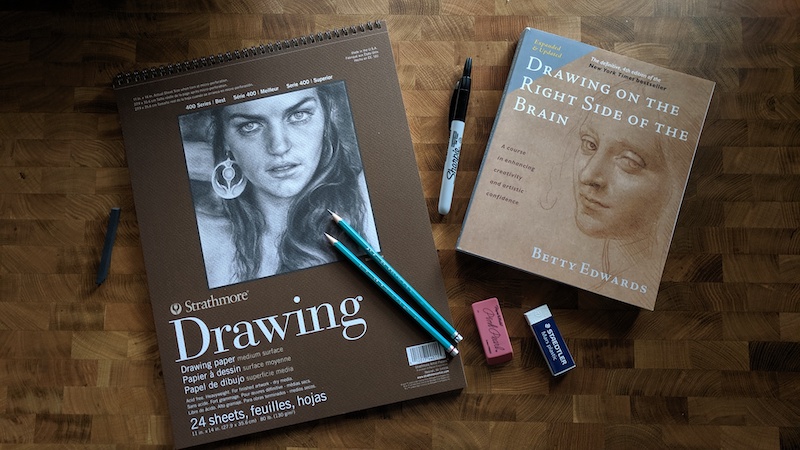 A spiral-bound pad of drawing paper with two drawing pencils, a marker, two erasers, and a textbook on drawing by Nic Rosenau on Unsplash.