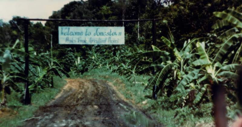 A sign hangs from wooden poles, reading Welcome to Jonestown Peoples Temple Agricultural Project. Trees and jungle are on both sides of the road. This photograph is located in the Moore Family's yellow album and was taken during John and Barbara Moore's May 1978 visit to Jonestown. A handwritten note on the back of the print reads, Entrance to Jonestown - one of two signs.
