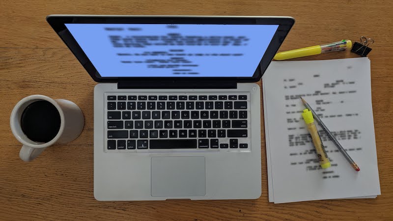 Flat lay photo of coffee cup, MacBook Pro laptop with script onscreen, printout of script, highlighter, pencil, pen, and binder clip by Nic Rosenau.