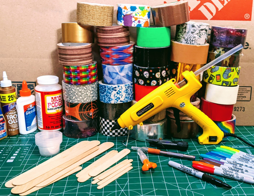 An assortment of glues, duct tape, mixing cups, crafting sticks, hobby knives, and markers with a hot glue gun. All in front of cardboard box and resting on a large green cutting mat. 2022. Photo by Nic Rosenau, CC BY-SA 4.0.