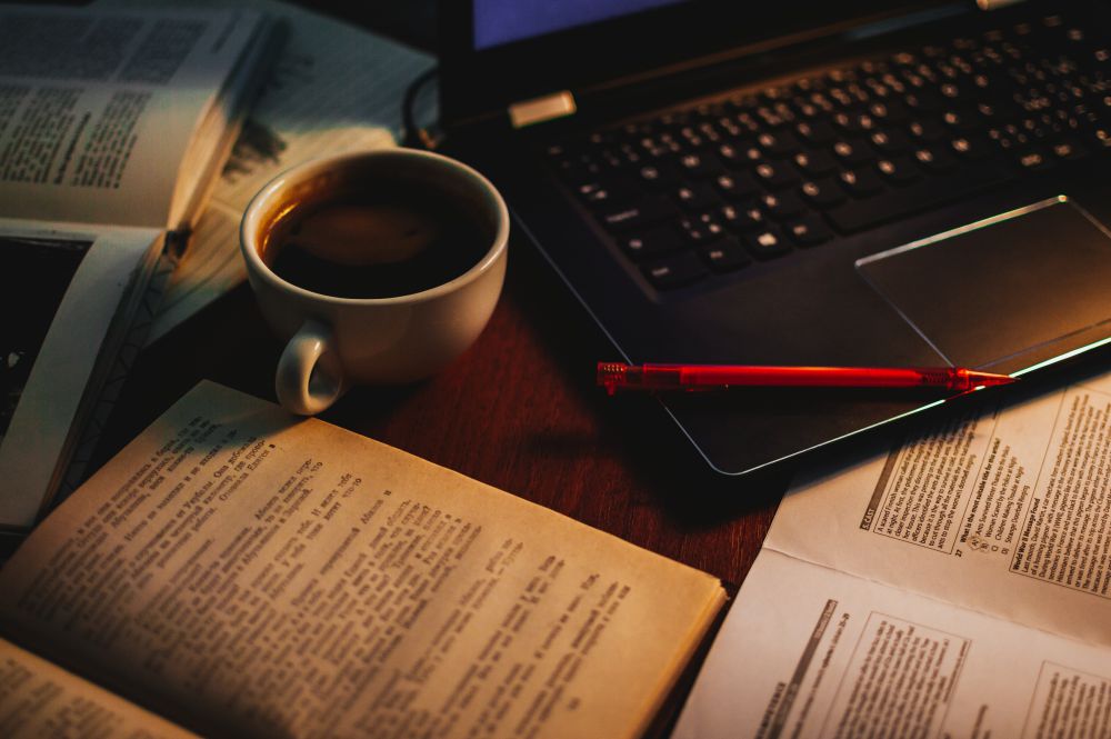 Photo of a cup of coffee and a red pencil in between open books, a notepad, and a black laptop, Prague, Czech Republic, 2020, by Vladyslav Bahara on Unsplash.