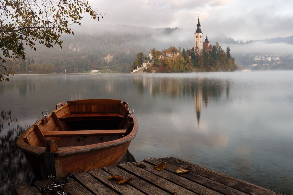 A foggy early morning at Lake Bled, Slovenia, with a rowboat at the dock in the foreground and Bled Island's Church of the Mother of God on the Lake in the background.