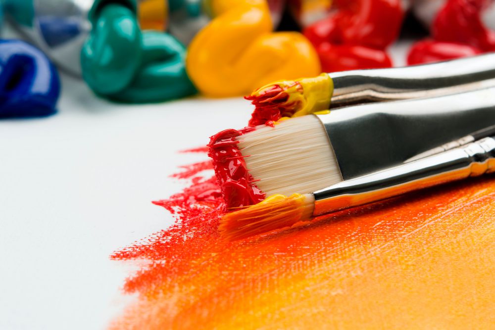 Three silver paintbrushes, one with yellow paint, one with red, and one with a mix of yellow and red. The brushes rest on a white canvas painted with a blend of red to orange to yellow. Dollops of blue, green, yellow, and red paint in the background.