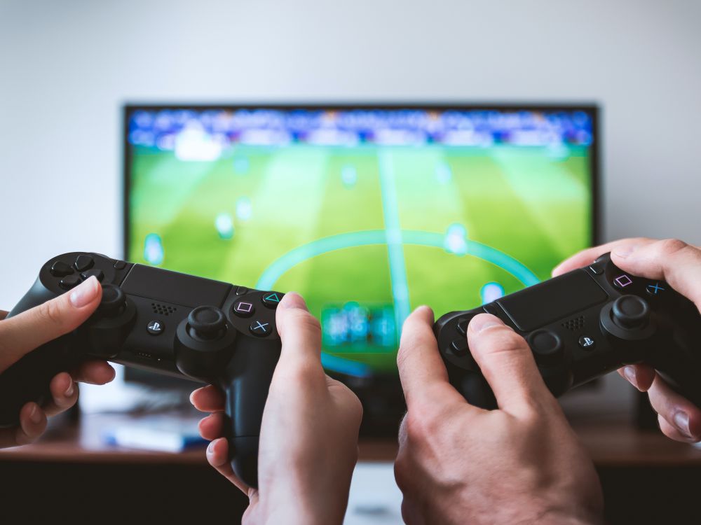 Two pairs of hands holding game controllers with a video game on a screen in the background. 2017. Photo by JESHOOTS.COM on Unsplash.