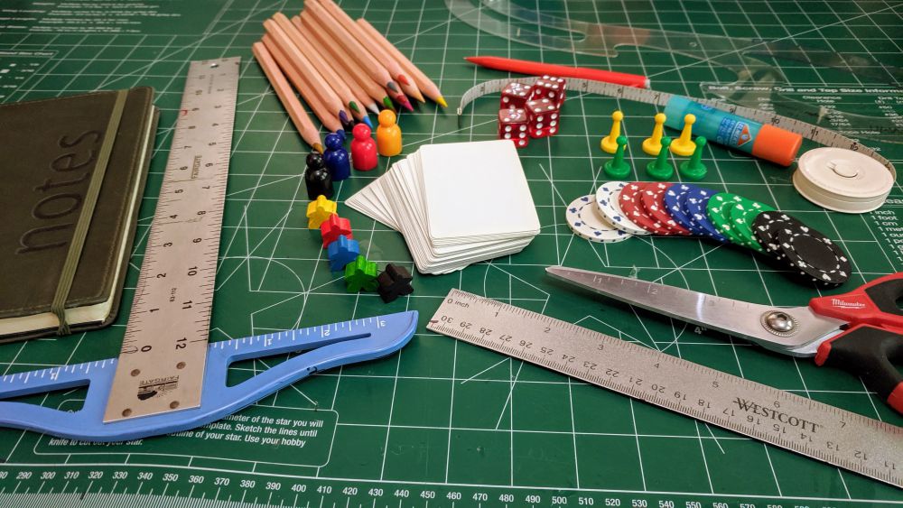 Homeschool board game design class supplies on a green cutting mat. Notebook, T-square, ruler, French curve, scissors, tape measure, glue stick, colored pencils, mechanical pencil, blank playing cards, dice, chips, pawns, meeple, poker chips. 2019. By Nic Rosenau.