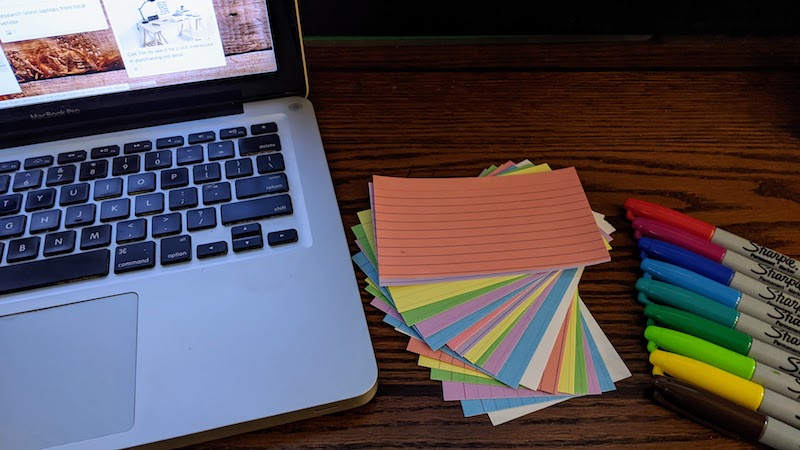 Brainstorming preparation. Collection of different colored Sharpie permanent markers next to a fanned-out stack of different colored index cards next to a silver MacBook Pro laptop resting on a dark wood desktop with a black soundbar on the back edge. Onscreen is the Ultimate To-Do List Trello Board Template by YouTuber Scott Friesen.