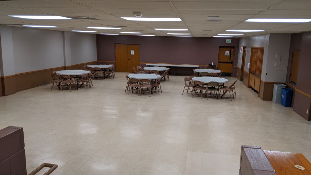 a large room with an open area and six five-foot round folding tables surrounded by 8 fold chairs each in the background