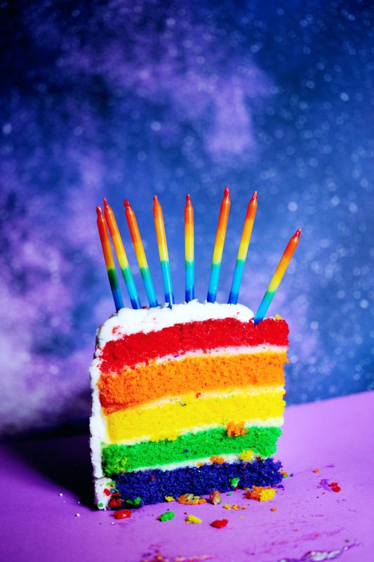 A colorful slice of rainbow layer cake with candles.