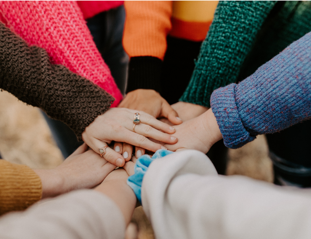 Eight hands piled together with arms sleeved by a variety of colorful sweaters. 2020. Decatur, AL, USA. 2020. Photo by Hannah Busing on Unsplash.